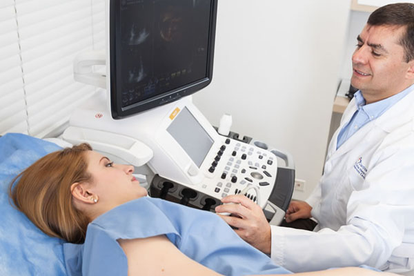Mujer con bata azul recostada mientras un doctor le hace un ecocardiograma mediante una herramienta